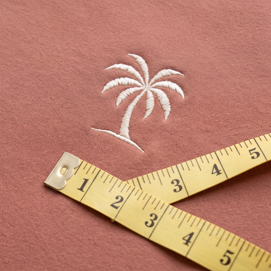 A yellow tailor's measuring tape lying across dusty mocha brushed cotton fleece below the cream embroidered feel it palm tree logo