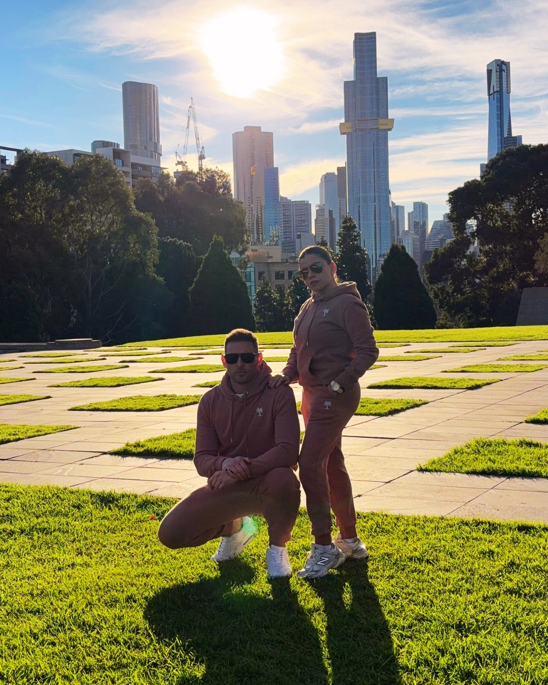 A man and a woman wearing matching mauve-brown activewear sets pose together on a sunny day in a city park with a skyscraper skyline in the background.